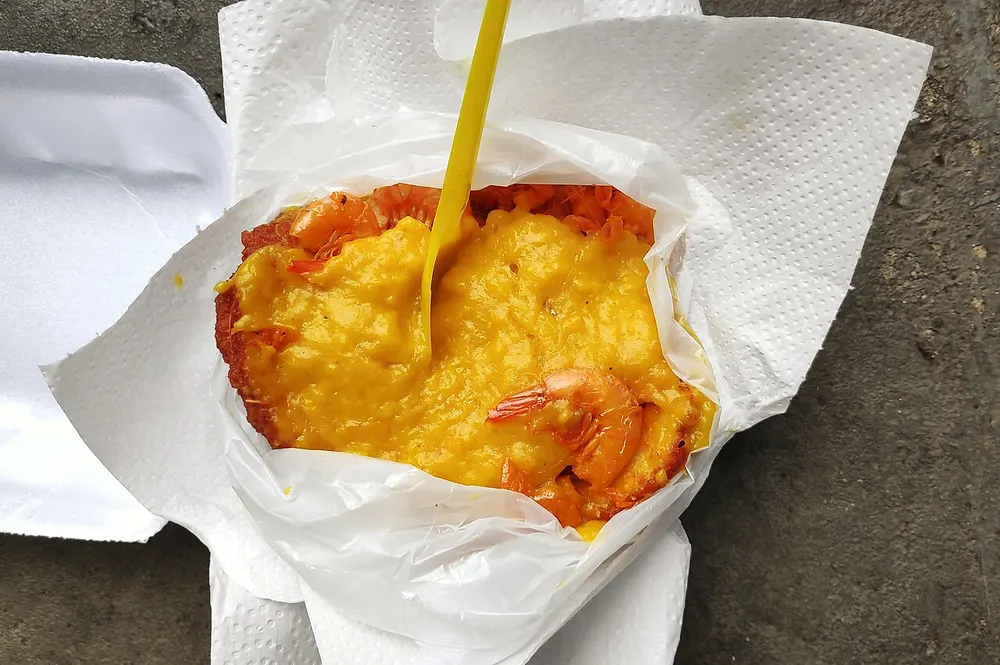 An acarajé split open in its street-stall wrapper — the deep-fried, red-brown bean fritter loaded with thick yellow vatapá and whole shrimp, a plastic fork stuck upright in it