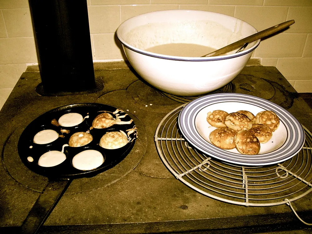Æbleskiver cooking in a dimpled iron pan on a wood stove, with the batter bowl and a finished plateful beside