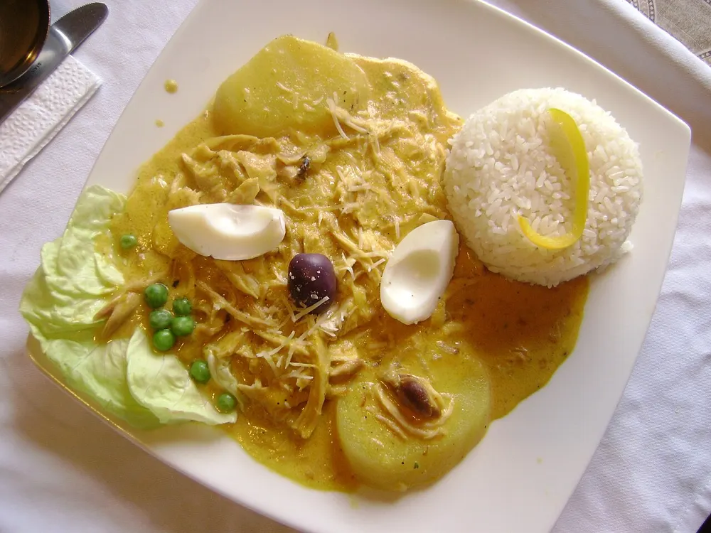 Ají de gallina over sliced potato with rice, egg, an olive and peas around it