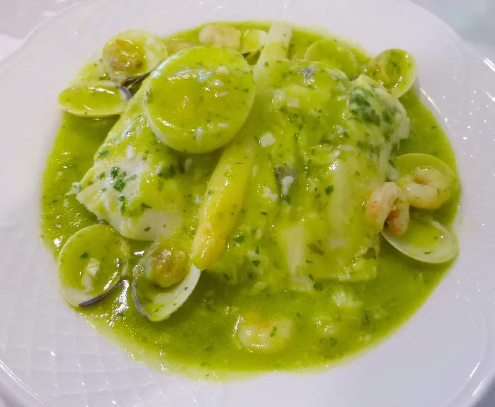 Hake fillets under a bright green parsley sauce, surrounded by open clams and white asparagus tips on a white plate