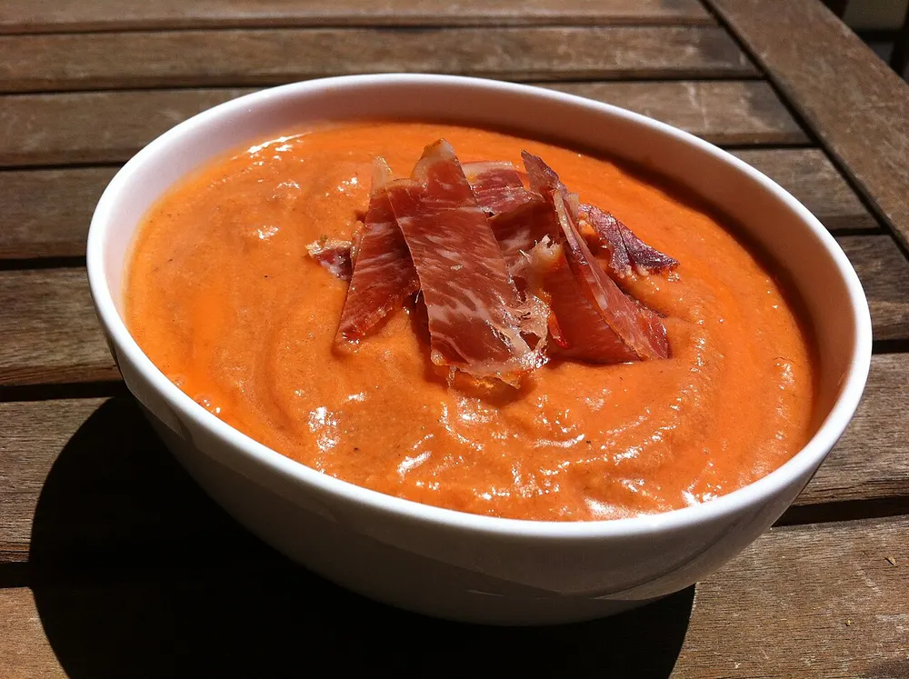 A white bowl of thick coral-orange salmorejo topped with strips of jamón ibérico, on a wooden table.