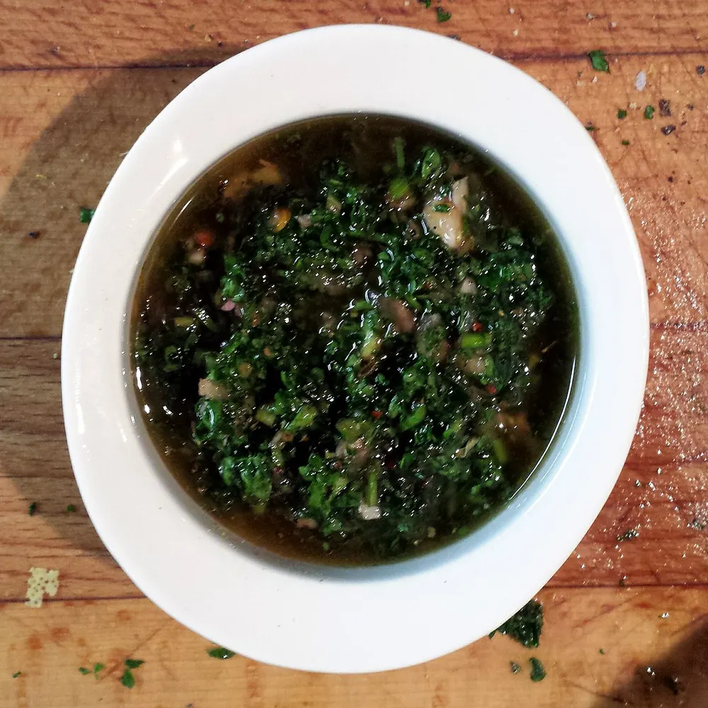 A bowl of green chimichurri, parsley and oregano loose in oil and vinegar with chili flakes