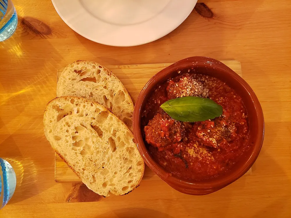 Meatballs in red tomato sauce in a terracotta dish, topped with grated cheese and a basil leaf, with two slices of bread beside it