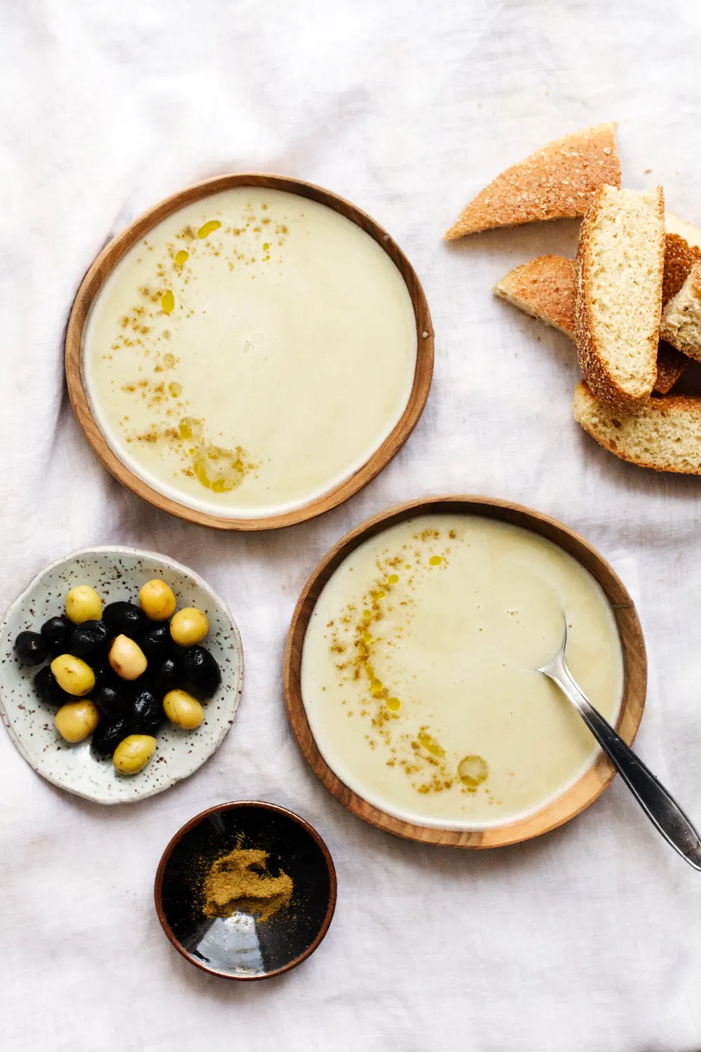 Two shallow bowls of bissara — smooth pale fava-bean purée drizzled with olive oil and dusted with ground cumin — set out with torn bread, olives and a dish of cumin
