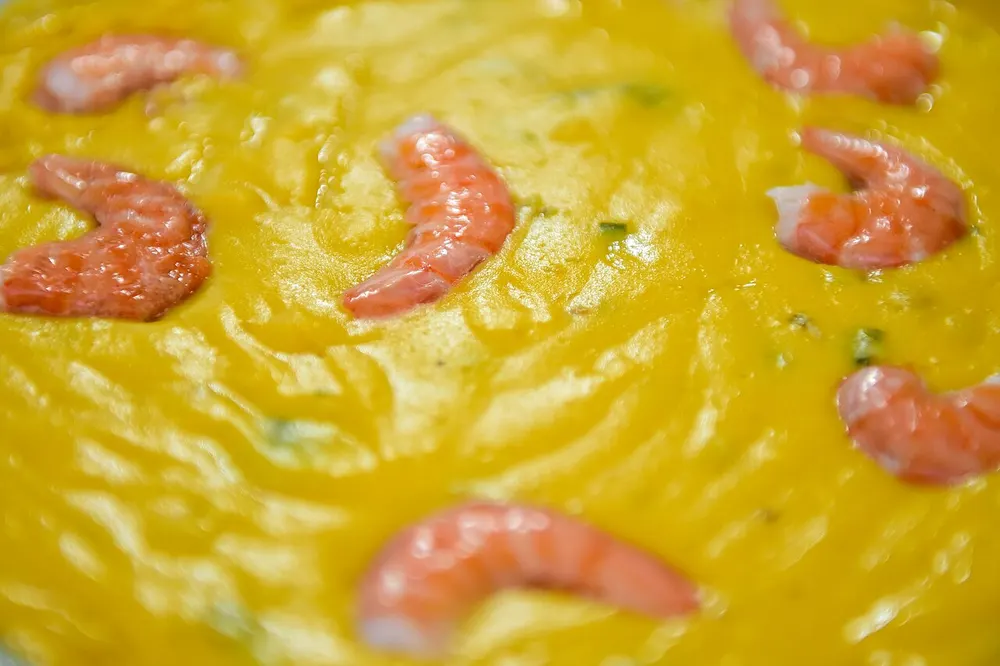Close-up of bobó de camarão — whole peeled pink shrimp resting in a thick, glossy golden-yellow cassava and coconut purée flecked with chopped coriander