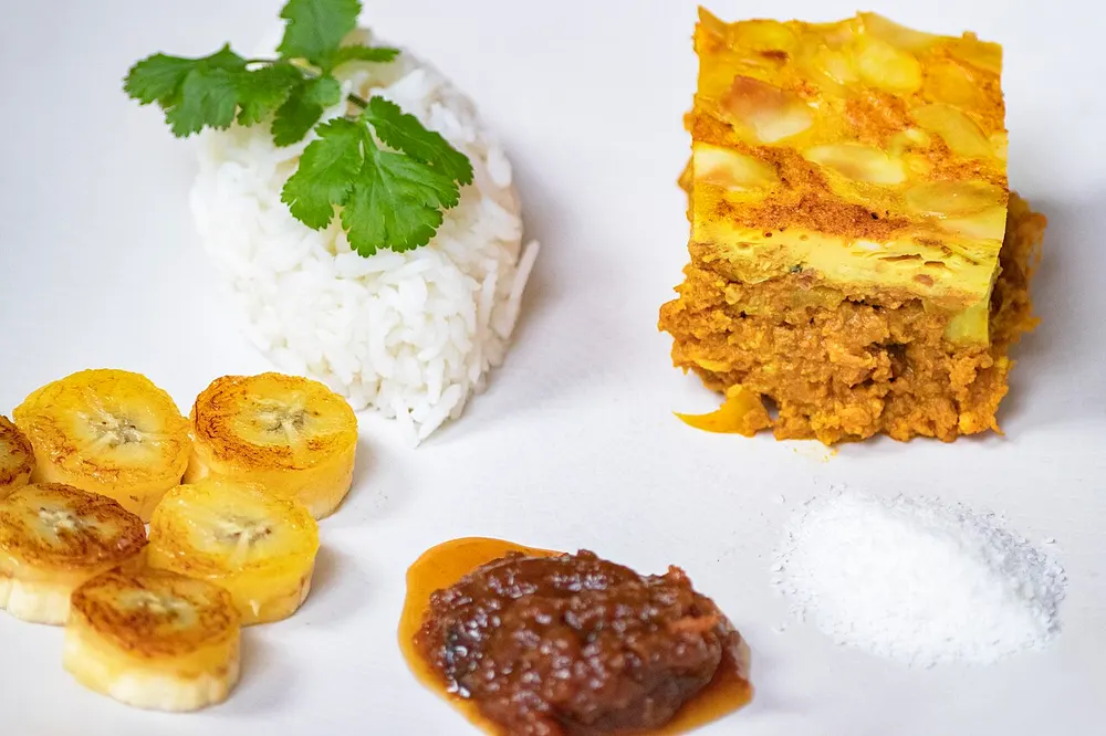 A cut square of bobotie showing curried mince under a set golden egg custard, with rice and chutney