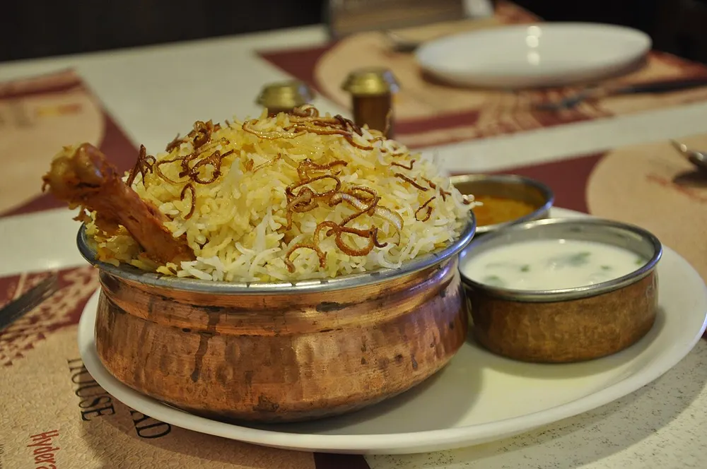 Saffron-streaked basmati biryani in a copper pot with crisp fried onions and a piece of bone-in chicken