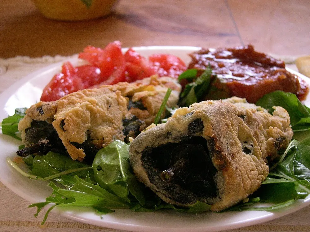Two battered and fried chiles rellenos on a white plate with greens, tomato and salsa