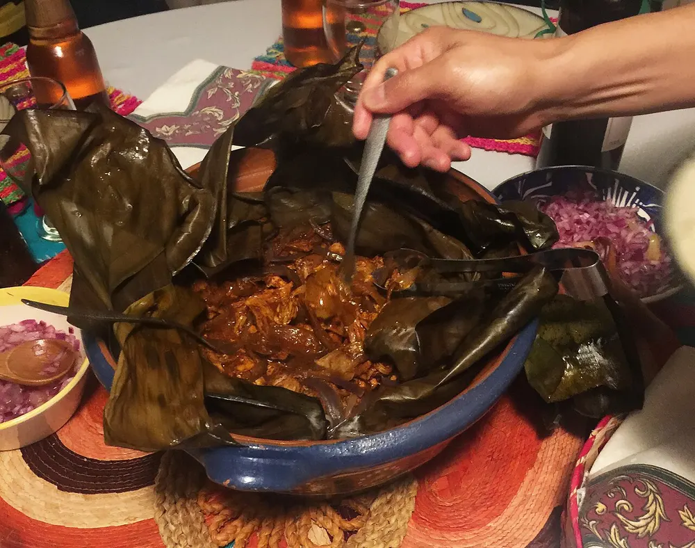 Shredded orange cochinita pibil in a blue clay dish lined with banana leaves, with pickled red onion nearby