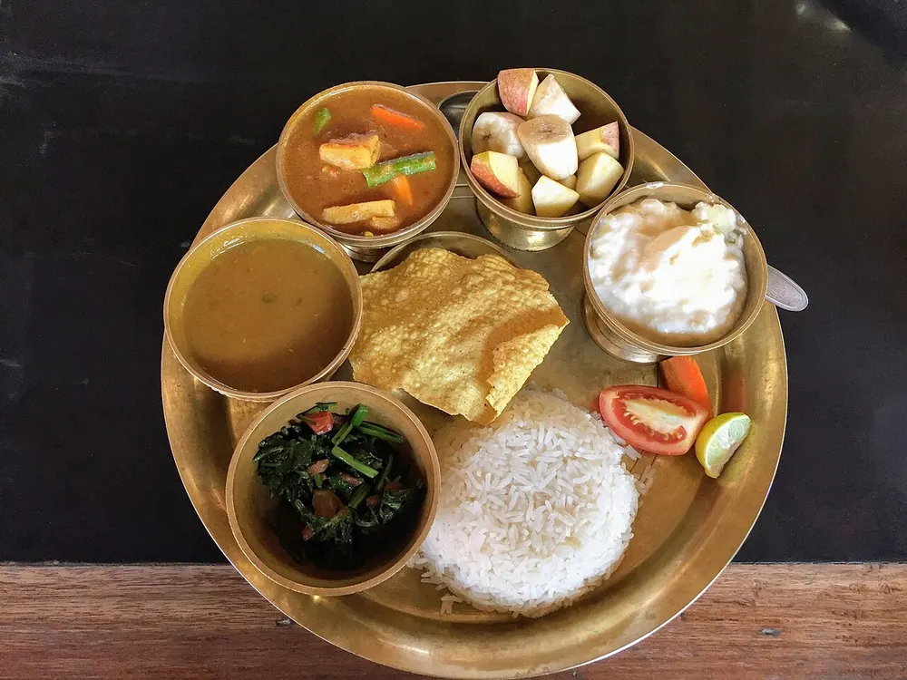 A dal bhat thali on a brass plate: rice, a bowl of dal, a vegetable tarkari, saag, papad, curd and fruit