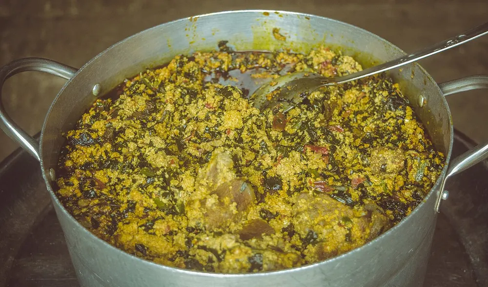 A large aluminium pot of egusi soup — pale yellow ground-melon-seed curds broken up through dark cooked greens and chunks of meat, with reddish palm oil pooling around the edge