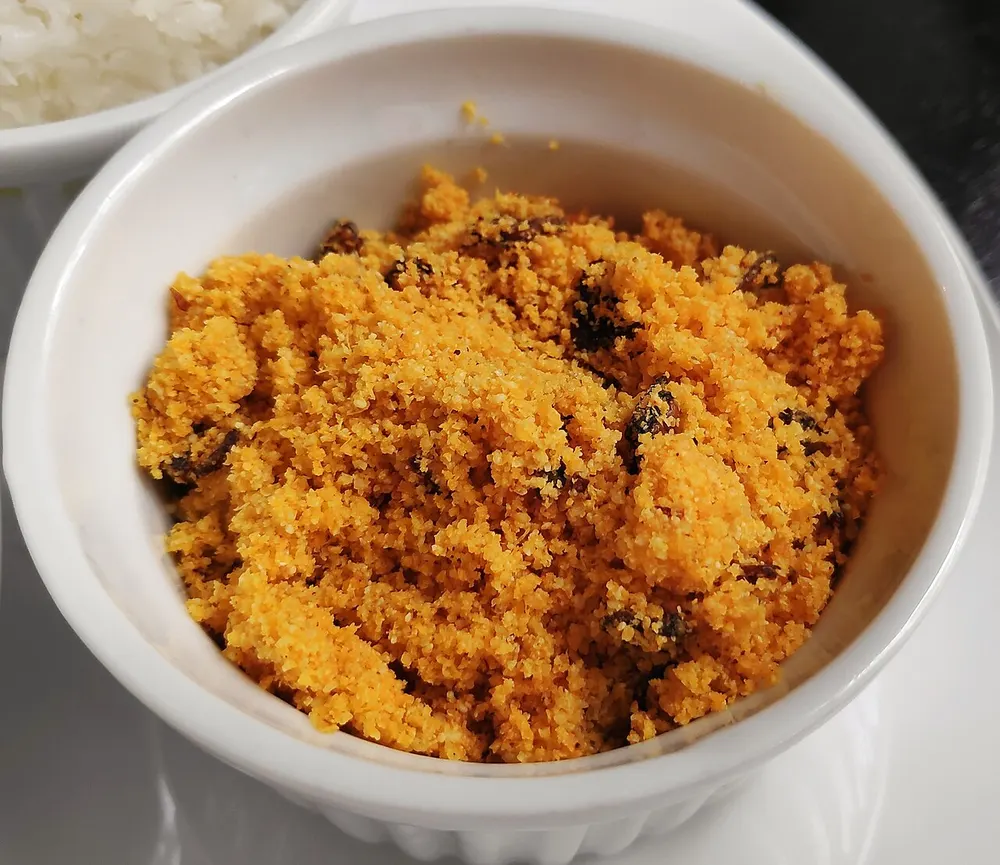 A small white bowl of farofa — cassava flour toasted to a loose golden-orange crumb with pieces of deeply browned onion scattered through it, a bowl of white rice behind