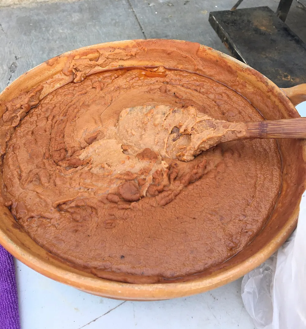 A wide clay dish filled with frijoles refritos — pinto beans mashed to a smooth, glossy brown purée, with a wooden spoon lifting a scoop from the middle