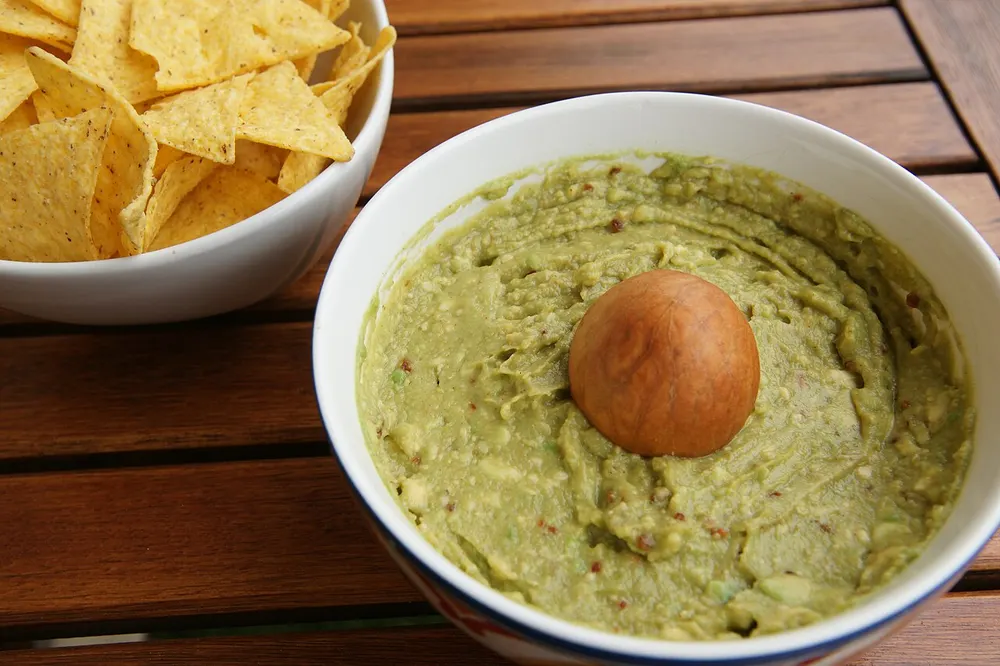 A bowl of mashed guacamole with the avocado stone set in the middle, tortilla chips alongside
