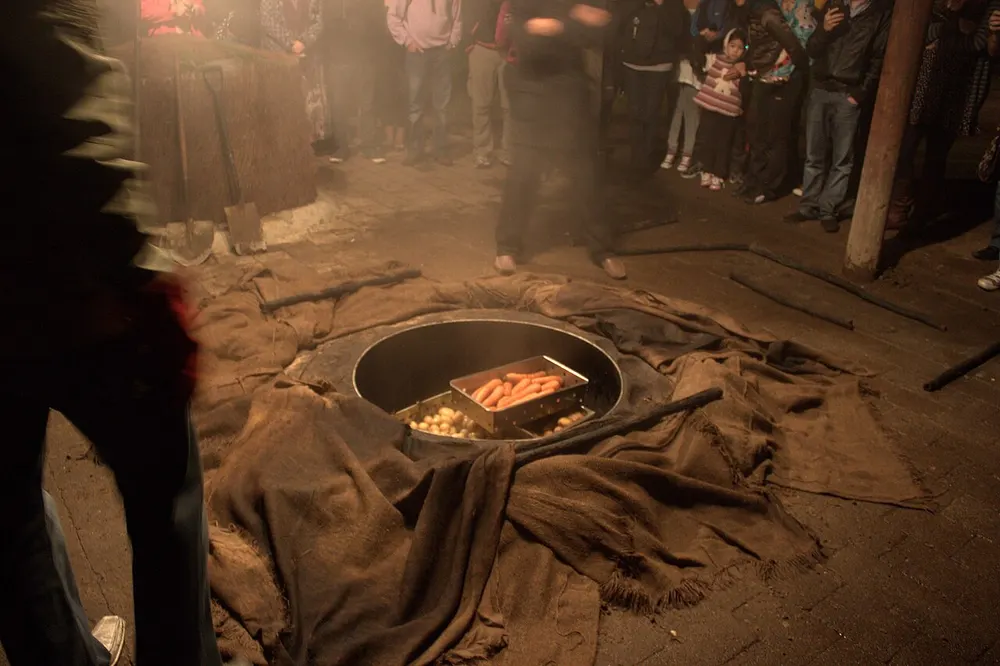 A hāngī pit uncovered at night — steam rising off wet sacking around the rim, a basket of potatoes and carrots down in the hole, and a crowd standing round it