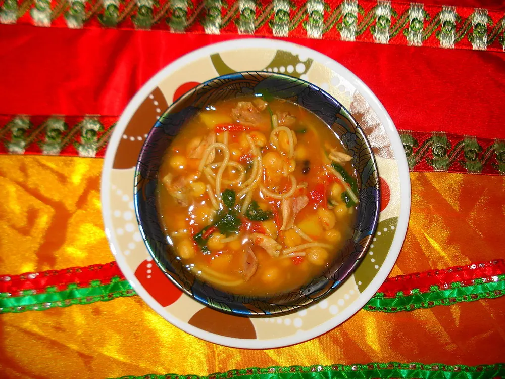 A bowl of harira seen from above — whole chickpeas, strands of vermicelli, shreds of braised meat and chopped coriander in a deep orange broth
