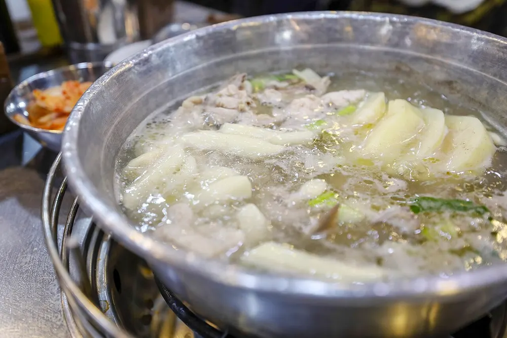 A wide metal pot of dak-hanmari bubbling on a burner — chicken pieces, rice cakes and green onion in a pale broth, kimchi alongside