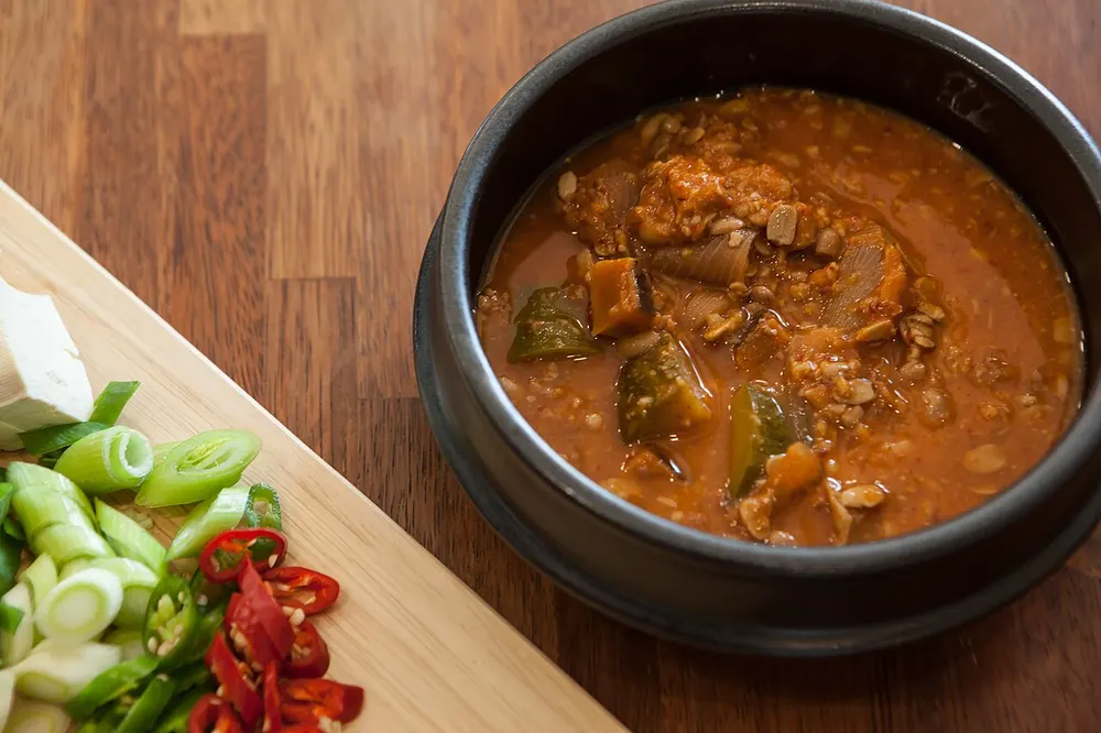 Doenjang-jjigae bubbling in a black earthenware ttukbaegi — a red-brown soybean-paste broth with chunks of zucchini and onion, beside sliced green onion and chilies on a board