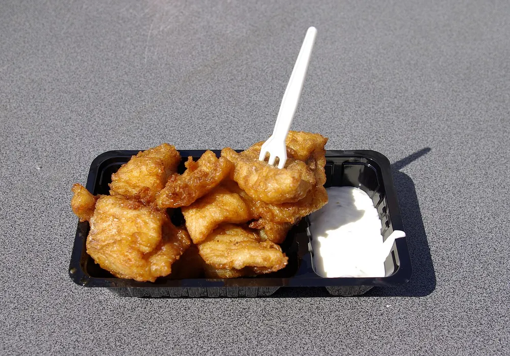 A tray of battered fried kibbeling with a scoop of white garlic sauce and a plastic fork