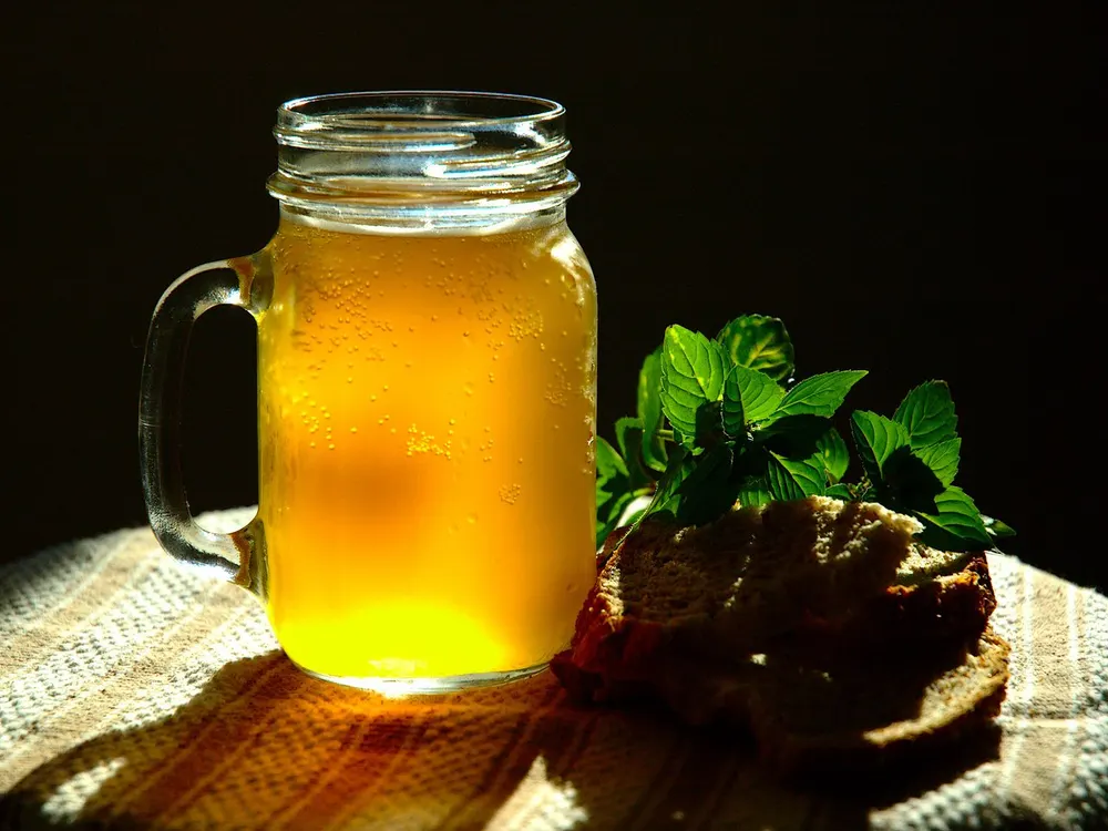 A jar of cloudy golden kvass with fresh mint and a slice of rye bread beside it