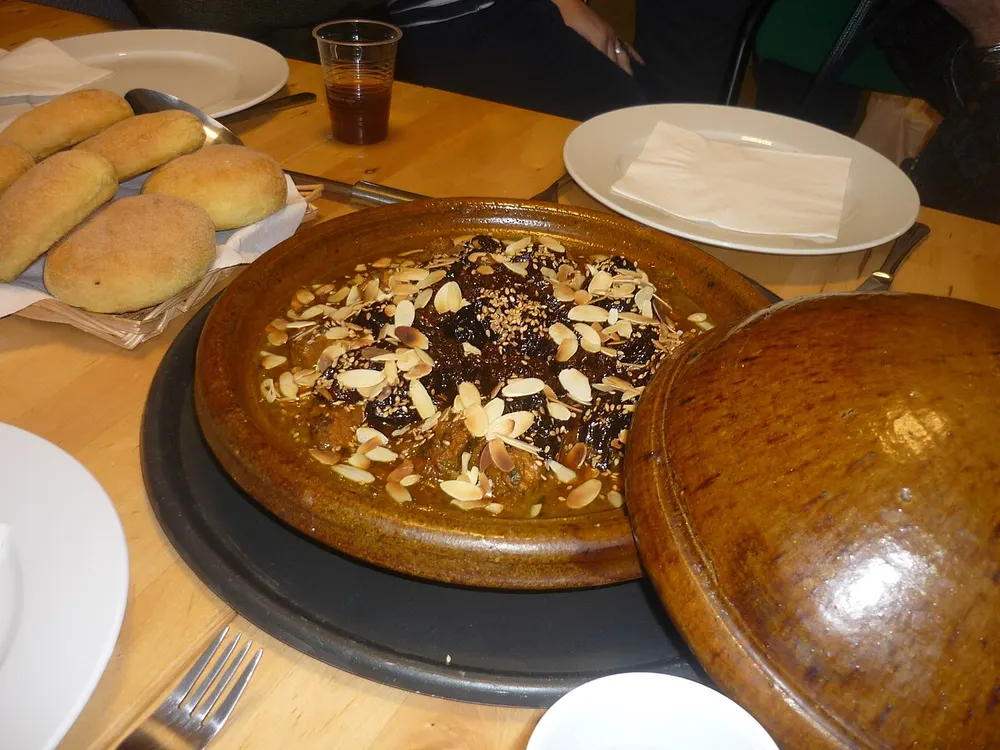 An open glazed clay tagine of braised meat in dark sauce, topped with glossy prunes, toasted flaked almonds and sesame seeds, its lid set to one side