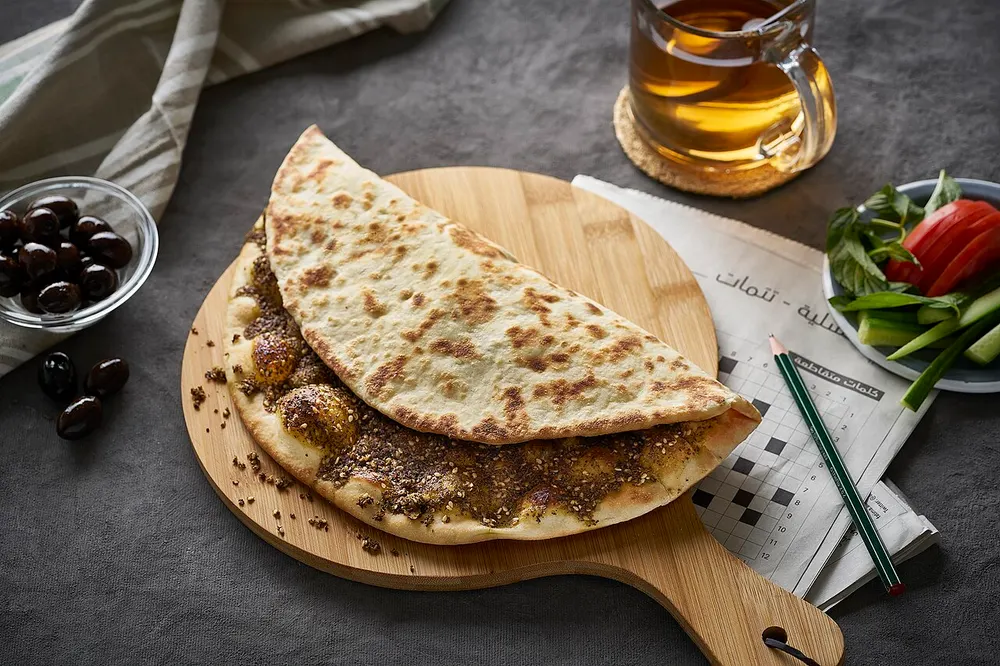 A za'atar manakish folded in half on a round wooden board — the dough blistered brown in patches, its underside spread edge to edge with dark green za'atar and sesame in olive oil, with olives and a glass of tea nearby