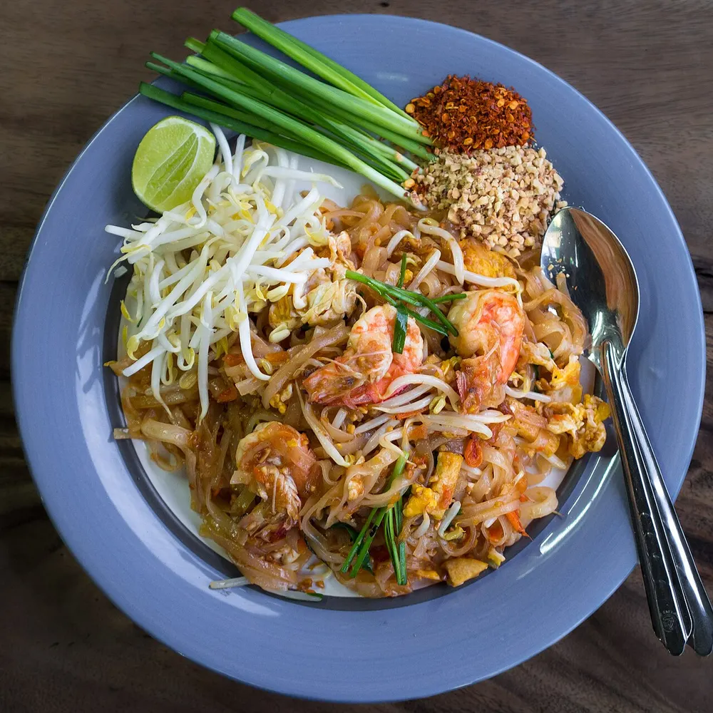 A plate of prawn pad thai with bean sprouts, chives, crushed peanuts, chilli powder and a lime wedge