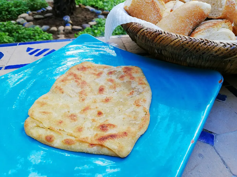 A single square-folded msemen on a turquoise plate, its surface blistered golden from the griddle and the folded layers visible along one edge