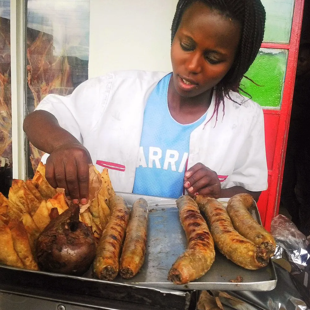 A Nairobi street vendor turning grilled mutura sausages on a tray beside samosas