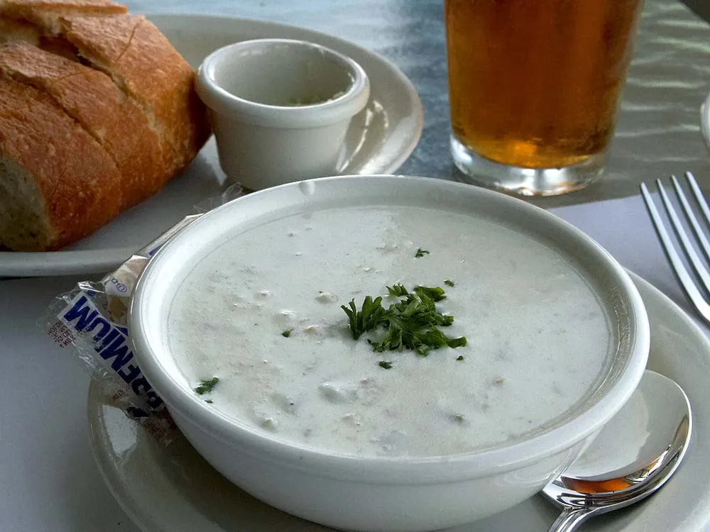 A white bowl of creamy New England clam chowder topped with parsley, with bread alongside