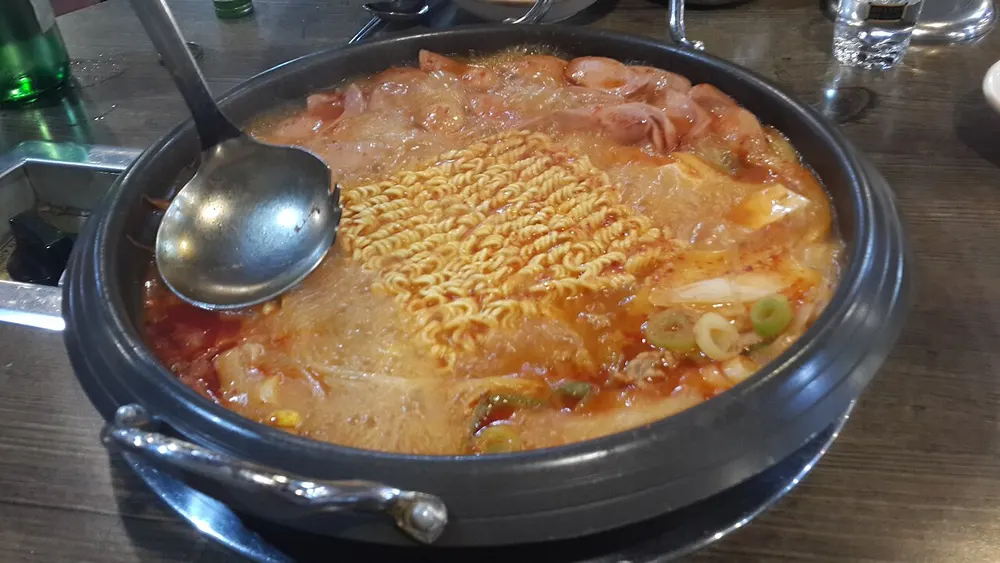A bubbling pot of budae-jjigae on a burner with sausage, ramyeon noodles and scallion in red broth