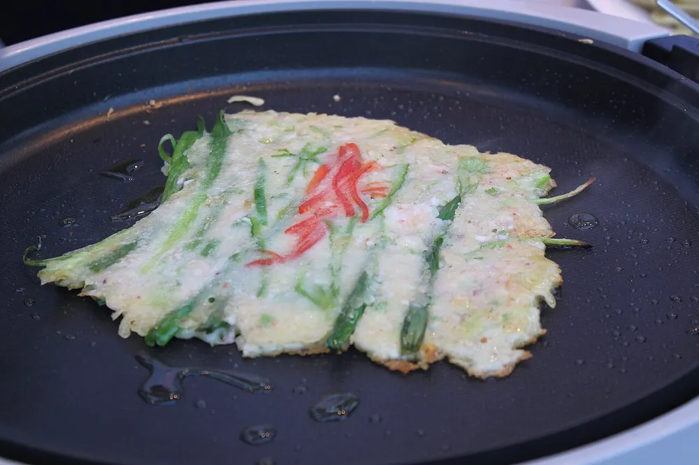 A scallion pancake cooking in an oiled pan, the green onions laid out in parallel with red chili slices on top