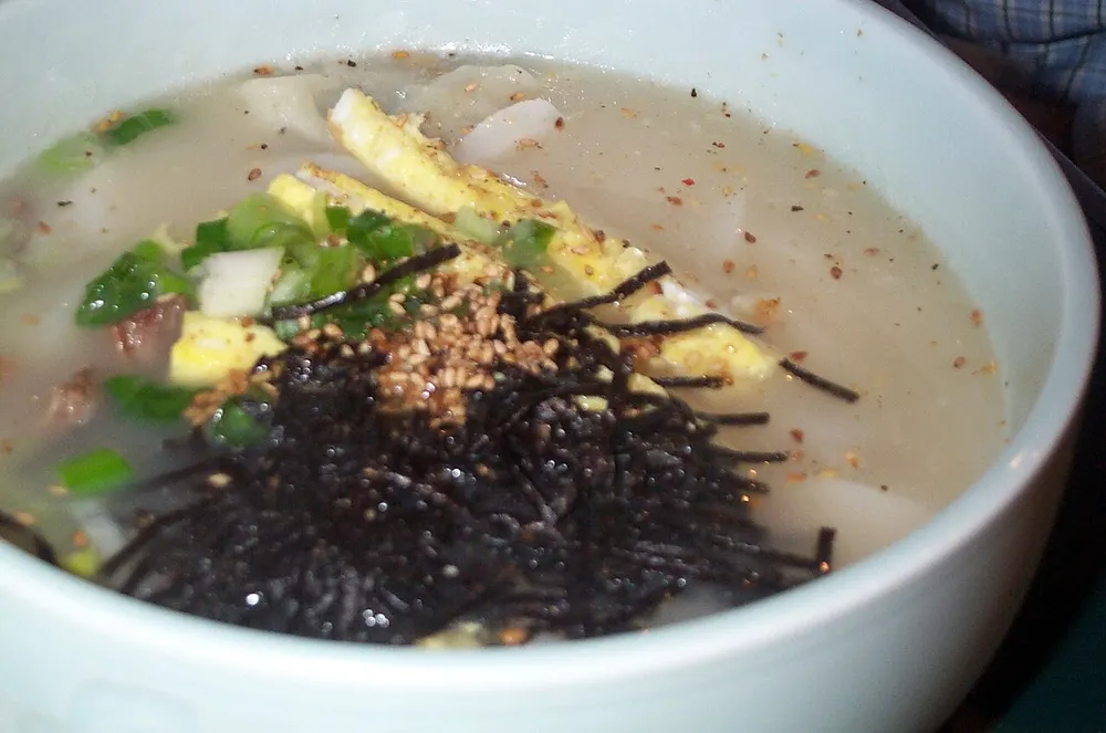 A bowl of tteok-manduguk — sliced rice cakes in milky broth topped with egg ribbons, seaweed flakes and scallion