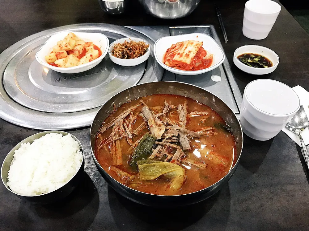 A pot of yukgaejang — red spicy soup with shredded beef and scallion — served with rice and banchan