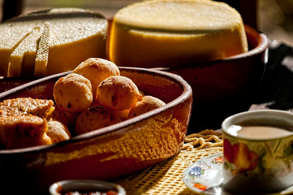 Golden pão de queijo in a clay dish, wheels of Minas cheese behind
