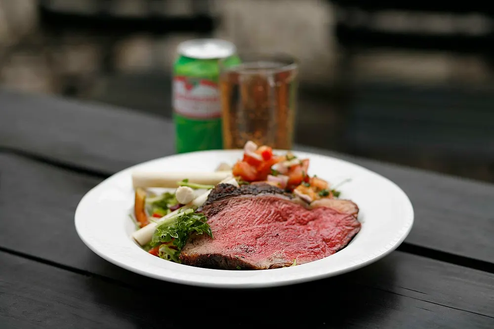 A thick slice of grilled picanha on a white plate — dark charred crust around the outside, rosy pink all the way through the middle, with a green salad behind it