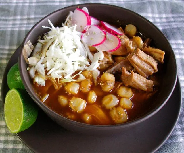A bowl of red pozole with hominy and shredded pork, topped with shredded cabbage and sliced radish, lime alongside