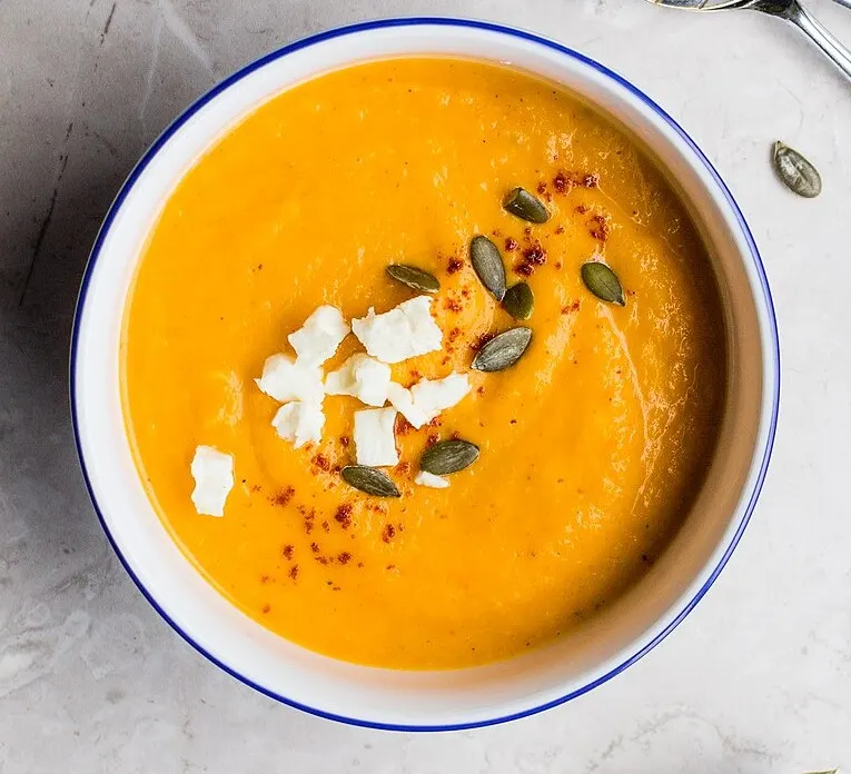 An overhead bowl of smooth, deep-orange pumpkin soup with a blue rim, finished with crumbled white cheese, green pumpkin seeds and a dusting of paprika