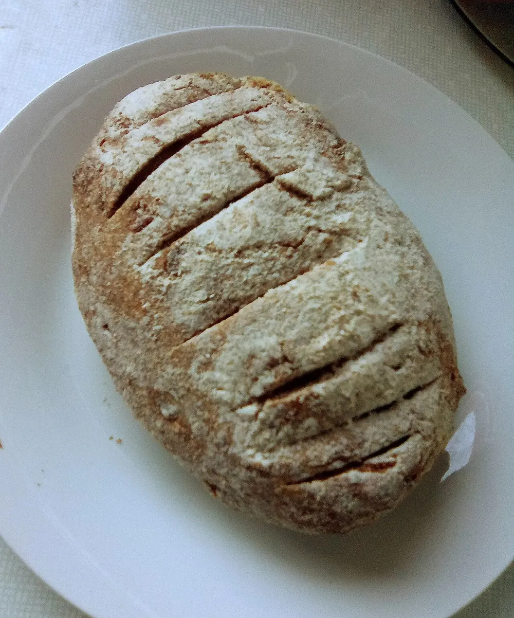 A round rēwena loaf on a plate, floured and slashed across the top, the crust cracked open along the cuts