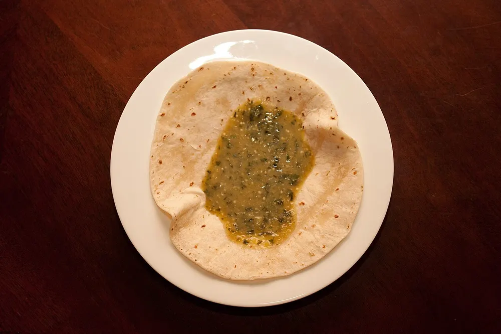A pool of salsa verde on a soft tortilla — olive-green tomatillo sauce with pale seeds and flecks of chopped coriander suspended through it