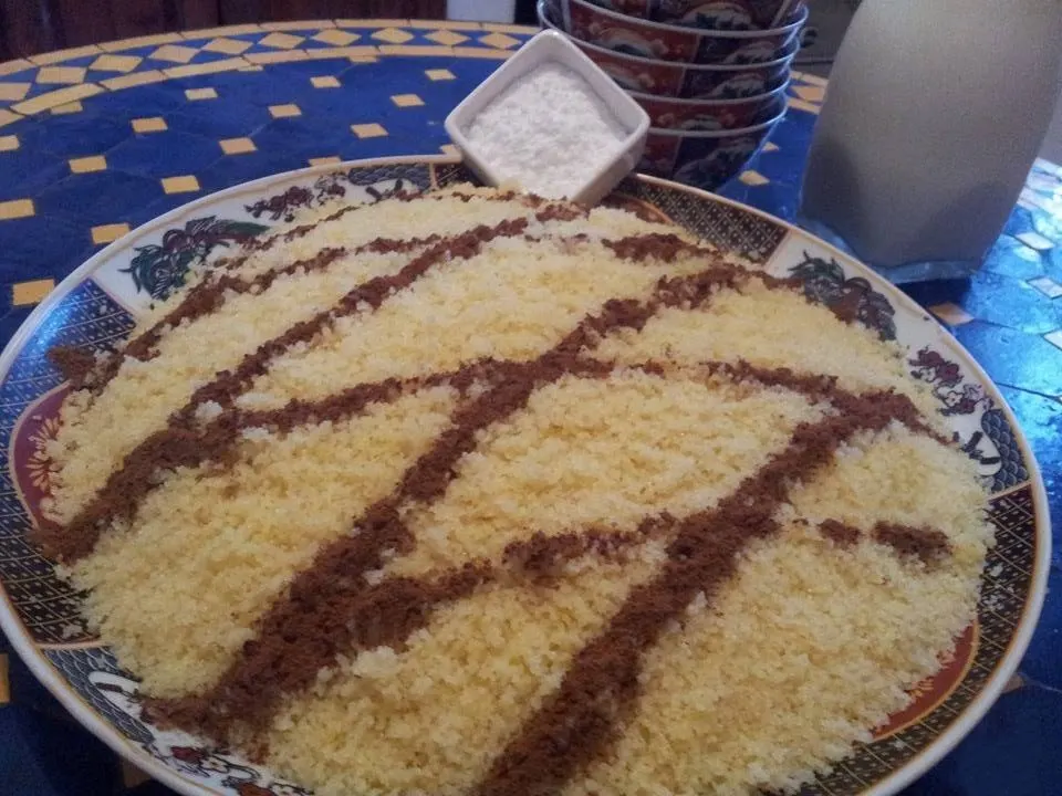 A wide decorated platter of steamed semolina seffa, mounded and combed into lattice stripes of ground cinnamon, with a small bowl of icing sugar beside it