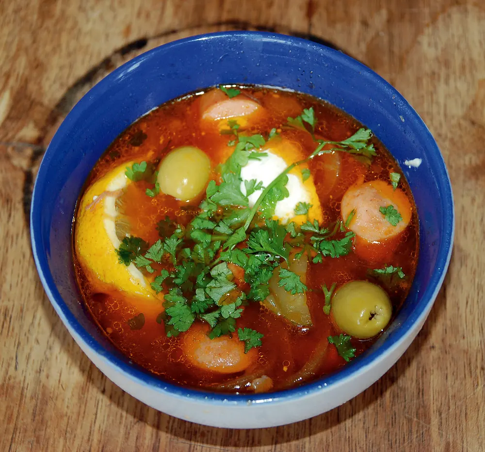 A bowl of solyanka with sausage, green olives, a slice of lemon, smetana and parsley
