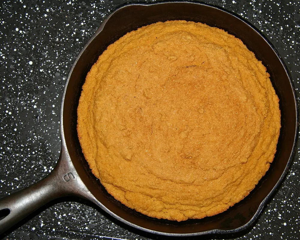A round golden-brown cornbread baked in a black cast-iron skillet, seen from above