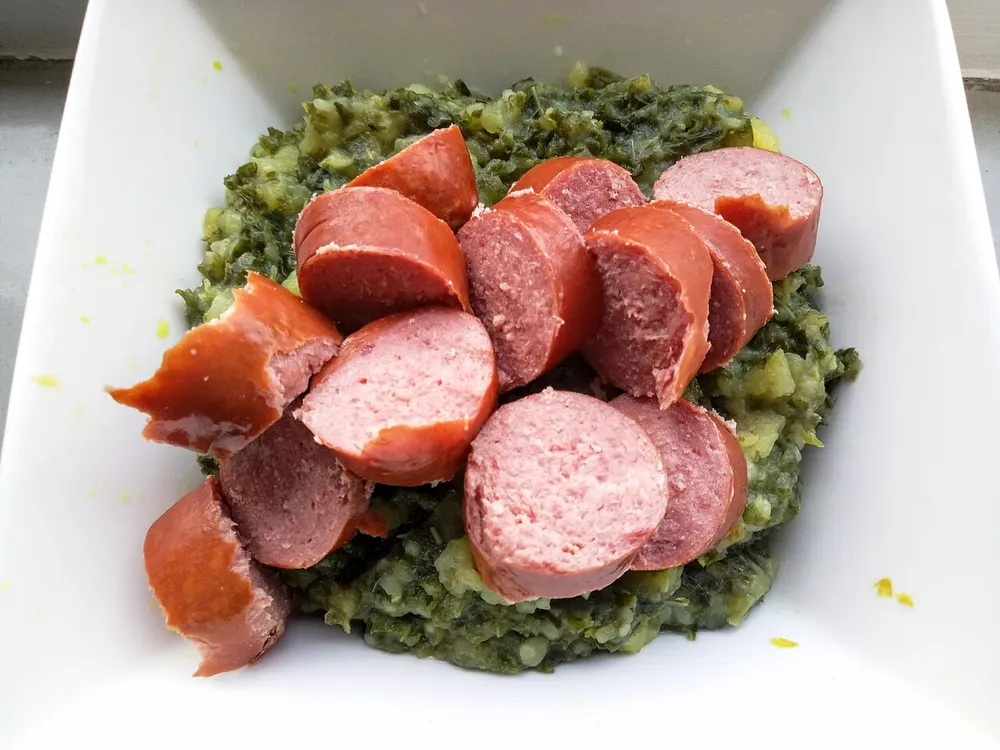 Kale stamppot in a white bowl with thick slices of rookworst laid over it