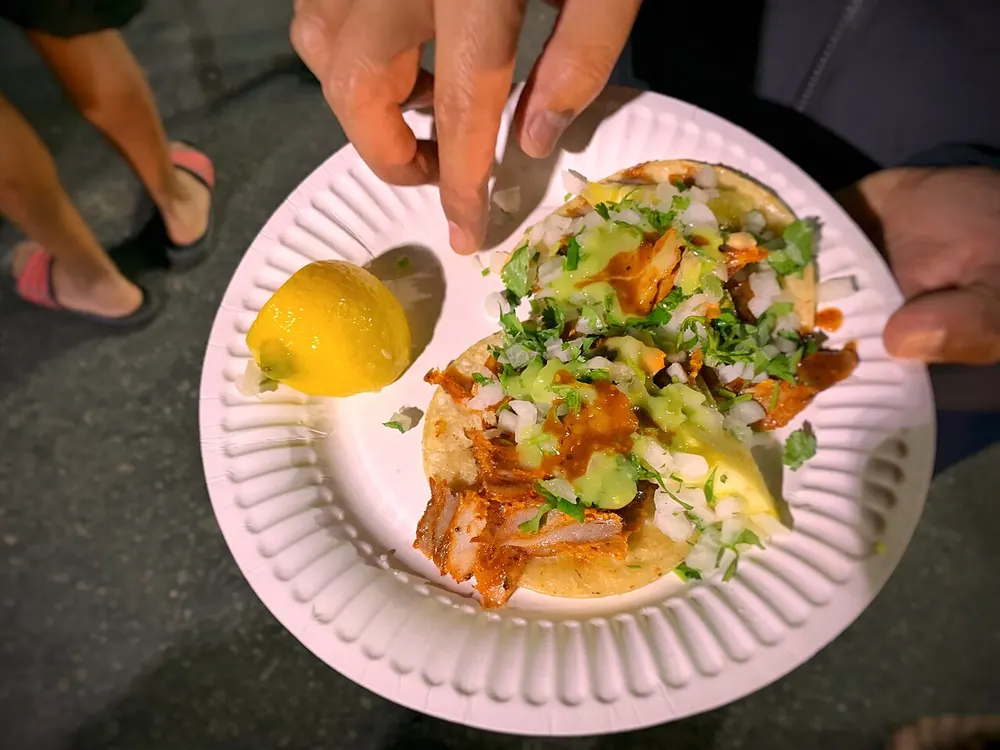 Two al pastor tacos on a paper plate with onion, cilantro and salsa, a citrus half alongside