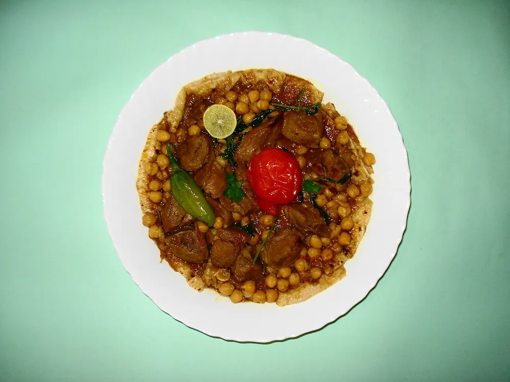 Tharid served flat — flatbread under lamb, chickpeas and sauce, with a lime and a green chilli on top