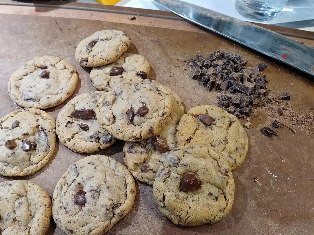 Chocolate chip cookies scattered on a wooden board beside a pile of hand-chopped chocolate