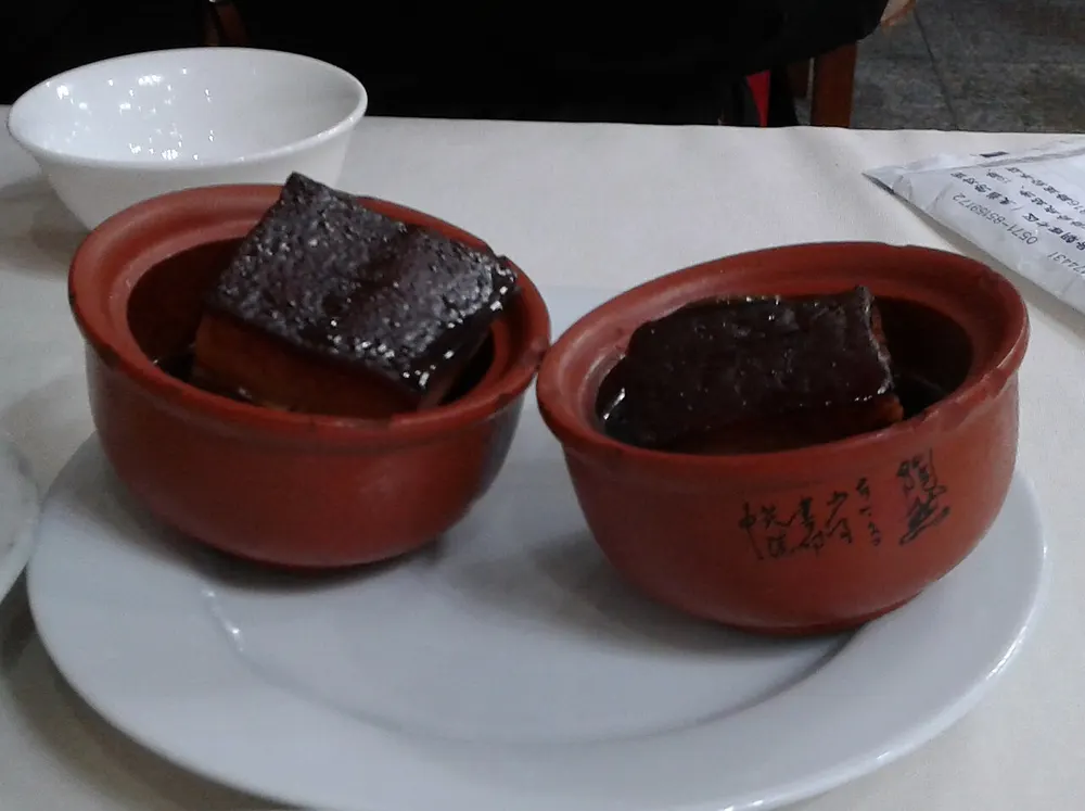 Two clay pots of Hangzhou-style dongpo pork, each holding a dark, glossy cube of braised pork belly