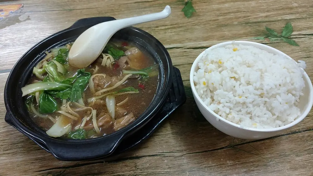 Huang men ji served in a black clay pot with greens and potato, beside a bowl of rice
