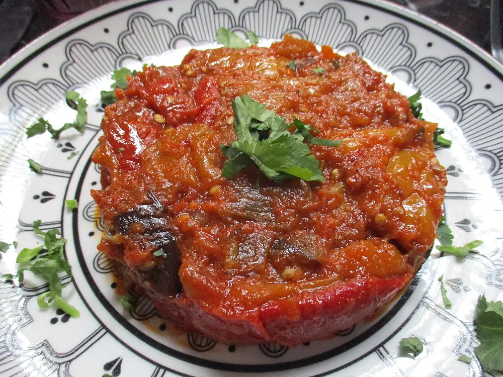 A round mound of zaalouk on a patterned plate — glossy brick-red cooked aubergine and tomato flecked with dark aubergine skin, topped with fresh coriander leaves